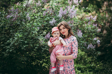 Obraz premium Young beautiful mother with small child in pink clothes on the background of beautiful blooming lilac in the open