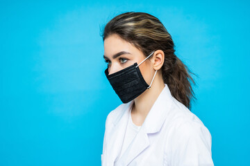 Young woman in a urgical suit, wears a protective mask on her face and gloves on hands posing while standing against a blue background. Protection. Care. Health. Nurse.