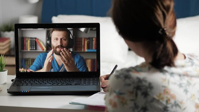 Doctor surgeon cardiologist online connection. Woman talking on internet video connection with man in blue medical suit and takes notes in notebook, doctor holds mock human heart in his hand