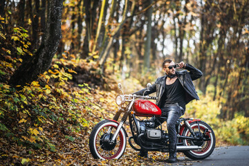 Plakat Bearded brutal man in sunglasses and leather jacket sitting on a motorcycle on the road in the forest