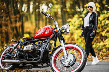 Pretty blonde biker girl in sunglasses with red motorcycle on the road in the forest