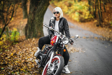 Plakat Pretty blonde biker girl in sunglasses with red motorcycle on the road in the forest