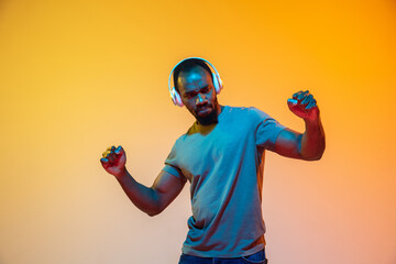 Dancing, delighted. African-american man's portrait in headphones on gradient orange background in neon light. Beautiful afro model. Concept of human emotions, facial expression, sales, ad. Copyspace.