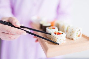 Female using chopsticks to pick up sushi roll from wooden plate. Japanese healthy traditional meal concept.