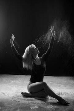 Beautiful plump blonde girl wearing a black gymnastic bodysuit covered with clouds of the flying white powder jumps dancing sitting on a dark. Artistic conceptual and advertising photo.