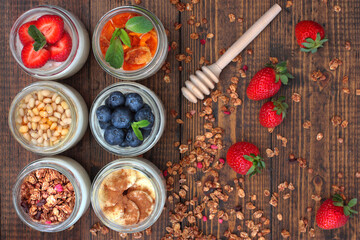 6 glass jars of home made yoghurt with different fillers: strawberries, blueberries, dried apricot, muesli, banana with cinnamon, cedar nuts with honey. Rustic wooden background witherries and muesli