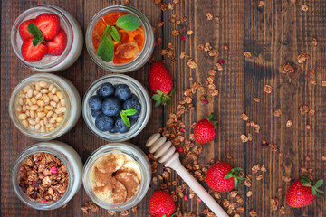 6 glass jars of home made yoghurt with different fillers: strawberries, blueberries, dried apricot, muesli, banana with cinnamon, cedar nuts with honey. Rustic wooden background witherries and muesli
