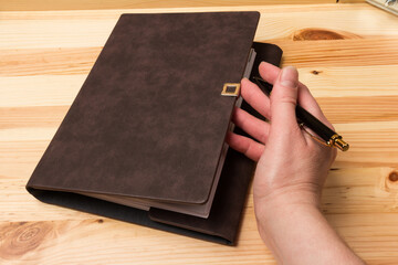 Leather-bound business notebook with a black pen in hand on wooden background. Business concept