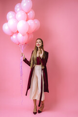 stylish young girl in burgundy clothes and pink balloons on a pink background smiles
