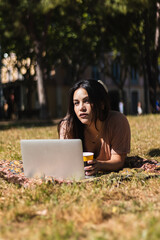 Hispanic woman lying on grass at park working on laptop and drinking coffee