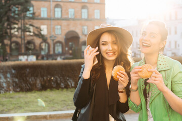 Positivity girls eating fast food, hamburgers, having dinner together, smiling at camera and posing. Beautiful couple of stylish girlfriends in leather jackets and hat, eating junk and unhealthy food.