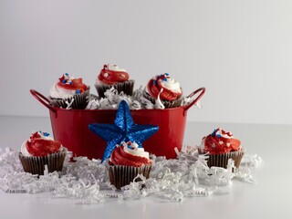 Mini chocolate cupcakes with red and white swirled frosting and star sprinkles with some in a red bucket with white paper shreds spilling out for 4th of July, Memorial Day, Veteran’s Day, Patriotic.