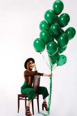 a young girl with a green pants and a black hat is sitting on a chair. Model posing with green bouncy balloons. celebrate concept
