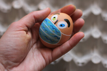 Painted Easter egg like human face with protective masks in the human hand, eggs carton on the background