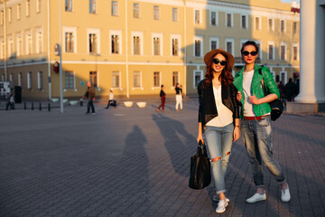 Two beautiful and shocked brunette girls stylish wearing walking at street, pointing by finger away and surprised looking with opened mouth. Emotionally girlfriends in sunglasses, hat, leather jacket.