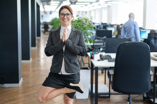 A pacified business woman doing yoga in an open space office. A red-haired smiling female employee in a casual suit and glasses stands in a tree pose at the workplace. Meditation and relaxation.