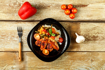 Baked chicken wings in tomato sauce with rice and vegetables on a black plate on a wooden table near vegetables for decoration.