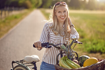 Woman using her bicycle to buy fresh produce standing on a rural road backlit by a warm glow of the sun smiling happily at the camera as she holds her bike with basket full of fruit and vegetables