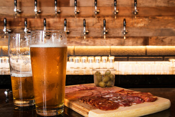 A cold glass of craft beer with a portion of snacks of cold cuts with olives, parma ham and salamis on wooden table over black background. Happy hour time.