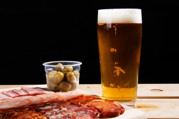 A cold glass of craft beer with a portion of snacks of cold cuts with olives, parma ham and salamis on wooden table over black background. Happy hour time.