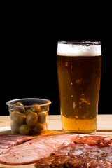 A cold glass of craft beer with a portion of snacks of cold cuts with olives, parma ham and salamis on wooden table over black background. Happy hour time.