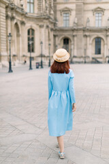 Fototapeta premium Back view of beautiful elegant young lady in hat and blue dress, walking near the vintage building in European city