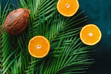Summer flat lay with palm leaf, oranges and coconut over dark green background.