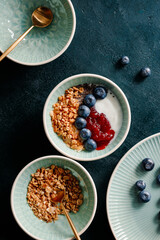 Top view of breakfast with granola, yoghurt, strawberry jam, chia seeds and blueberry on a dark green table.