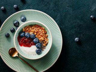 Top view of breakfast with granola, yoghurt, strawberry jam, chia seeds and blueberry on a dark green table.
