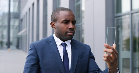 African American young businessman in suit and tie standing at street and holding piece of glass on which tapping and scrolling like on phone screen. Touchscreen future technology. Transparent device.