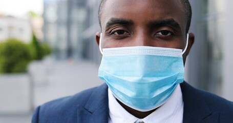 Close up of face of young African American handsome businessman in medical mask looking straight to camera with happy look. Portrait of man at street. Coronavirus concept.