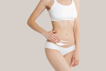 Fototapeta na wymiar Slim young woman in underwear apply cream to belly on beige background
