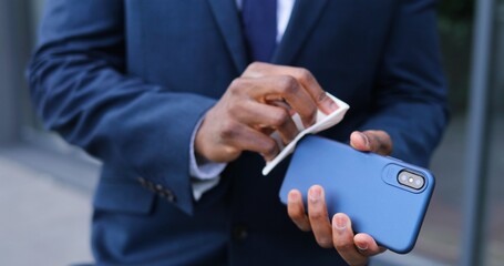 Close up of smartphone in hands of African American businessman in suit and tie. Man cleaning phone with disinfecting napkin. Cellphone disinfection outdoor.