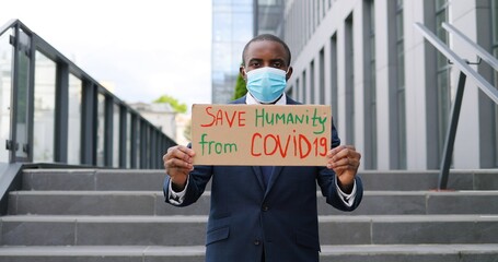 Portrait of carton poster with words Save Humanity from Covid 19 in hands of African American young man in medical mask. Lonely protest outdoors on town square. Male healthcare activist.
