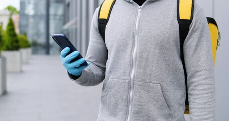 Close up of African American man in medical mask and gloves outdoor texting message on smartphone. Deliveryman tapping and typing on mobile phone, using map navigator and looking for address.