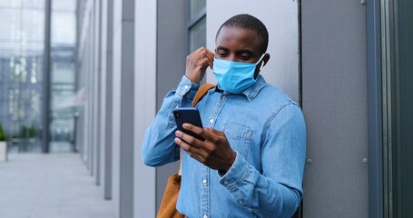 Fototapeta na wymiar African American man in medical mask standing on street and texting message on mobile phone. Male in respiratory protection is outdoors and tapping or scrolling on smartphone. Browsing online.