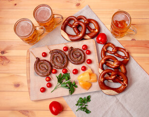 Cutting board with roasted sausages, beer mugs and soft pretzels on wooden table. Top view.