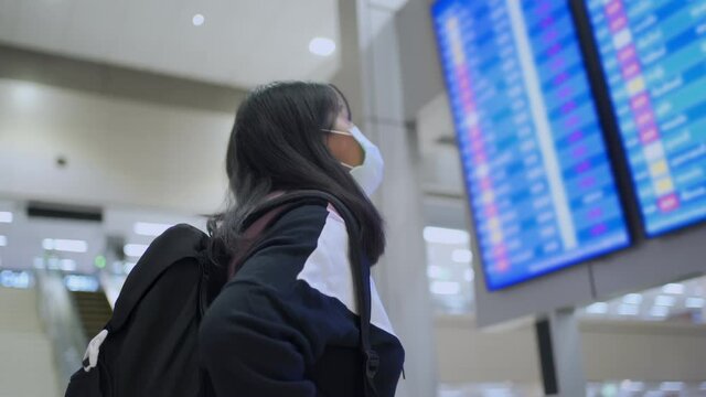 Asian female in white mask looking  up to airport flight time table board, at airport terminal,  corona virus covid-19 pandemic, new normal lifestyle social distance, blue led screen leaving city, 4k
