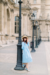 Obraz premium Full length portrait of beautiful young smiling Caucasian woman in elegant blue dress and hat, standing near the old vintage building and leaning on vintage street lamp