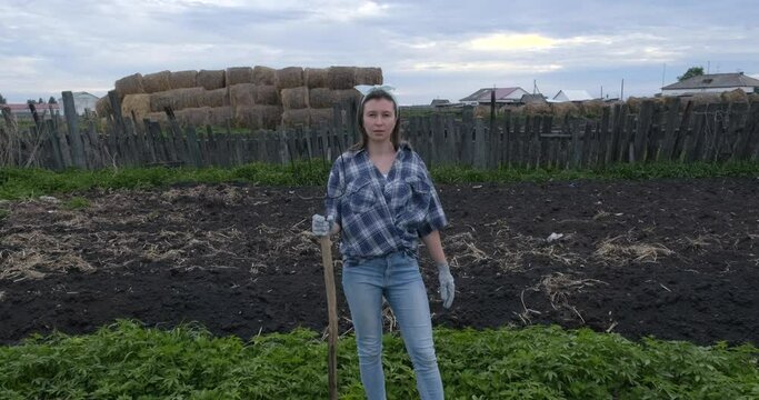 Girl Stands on Background of Plowed Land and Lopsided Wooden Village Fence. Holds Garden Tool. Pitchfork. Village. Resident of Village. Portrait. Posing. Simpleton. Hard Look. Insects curling around