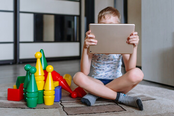 The boy is in the game room, next to toys, but does not play, but looks at the tablet