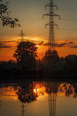in the mirrored surface of the pond, the power line tower is illuminated by the setting sun
