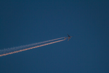 the plane is flying high in the blue sky and the setting sun beautifully illuminates the contrail