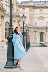 Side view of beautiful young woman in elegant blue dress and hat, standing near the vintage...