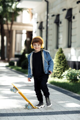 Photo of joyful little boy posing with headphones and skateboard in street.