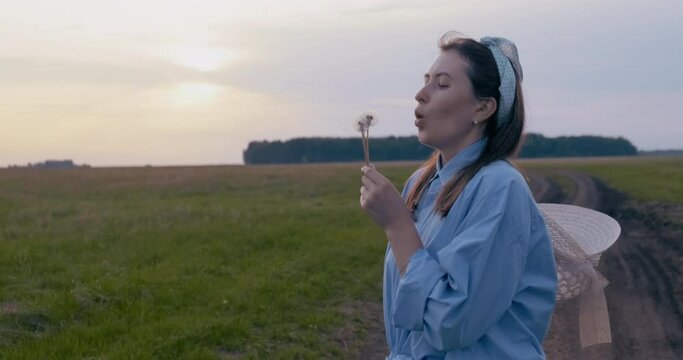Girl Stands in Field. Hands Holding Dandelions. Blowing on Flower. White Seeds Scatter Around. Beautiful Hillbilly. Turns to Camera. Smiles. Shy. Posing. Shows Emotions. Sunset. Freedom. Open space