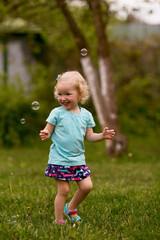 Little cheerful girl with blond hair enthusiastically plays with soap bubbles in the garden. Happy carefree childhood. Summer leisure for children.