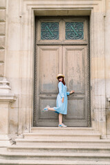 Fototapeta na wymiar Charming elegant young woman wearing blue dress, posing in front of ancient wooden door of old vintage building with one leg up