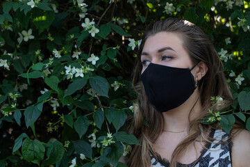 Pretty young blonde woman in medical black face mask. in a bush with little white flowers. modern reality. covid-19 concept. copy space. High quality photo