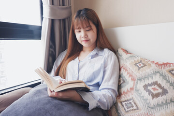woman reading book in bed next to a window.